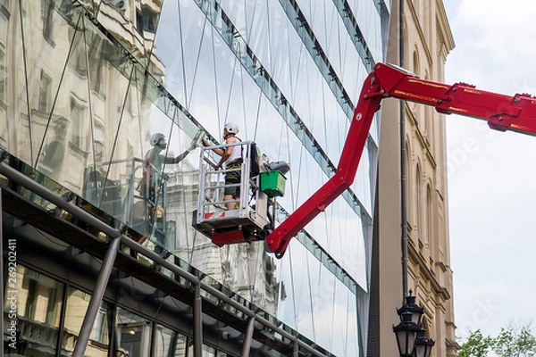 Fototapeta Worker in elevated work platform cleaning building facade window glass by using equipment tools to cleanup dirty surface. Selective focus