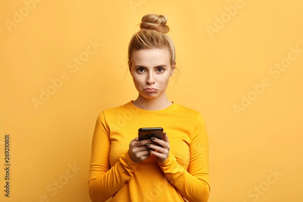 Fototapeta angry sad woman with hairbun annoyed by something while using phone, girl has received bad sms, text message isolated yellow background. emotion, reaction, feeling
