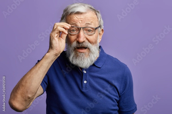 Fototapeta cheerful old man looking through the glasses at the camera isolated on blue background. close up portrait. studio shot.handsome man cannot recognize his grandson. man with poor eyesight.