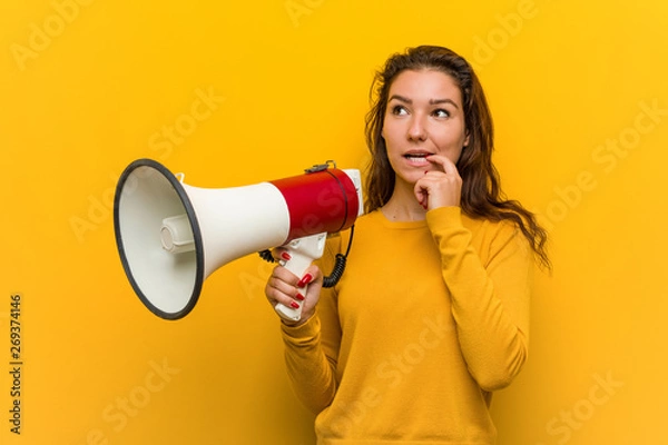 Obraz Young european woman holding a megaphone relaxed thinking about something looking at a copy space.