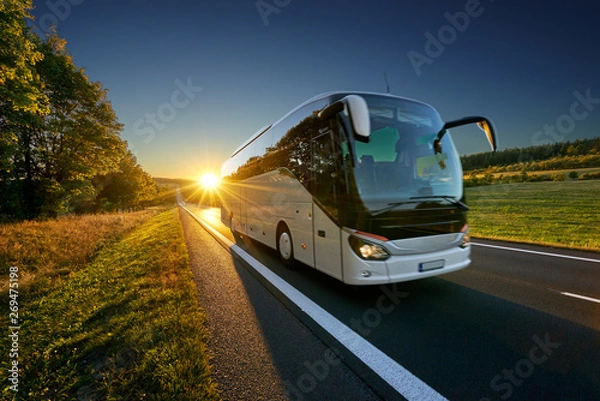 Fototapeta Biały autobus poruszający się po asfaltowej drodze wokół linii drzew w krajobrazie wiejskim o zachodzie słońca
