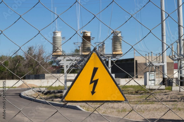 Obraz Electrical hazard sign on metal grid. On the background of unfocused high-voltage electrical substation. Concept: danger of electric current, death from electric shock