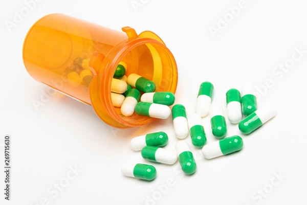 Fototapeta A bunch of green and white capsules spilling out of a slightly transparent orange medicine pharmacy bottle set on a plain white background in horizontal image format.