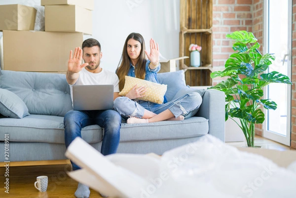 Fototapeta Young beautiful couple relaxing sitting on the sofa of new house using computer laptop with open hand doing stop sign with serious and confident expression, defense gesture