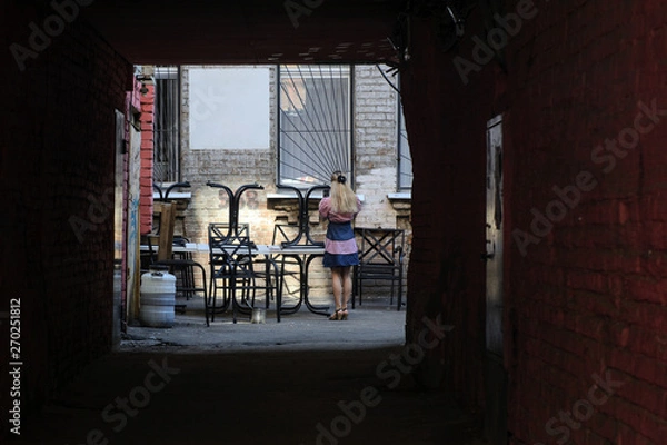 Fototapeta The passage to the old courtyard in the ancient city illuminated by the sun. Table, chairs and a girl with a phone