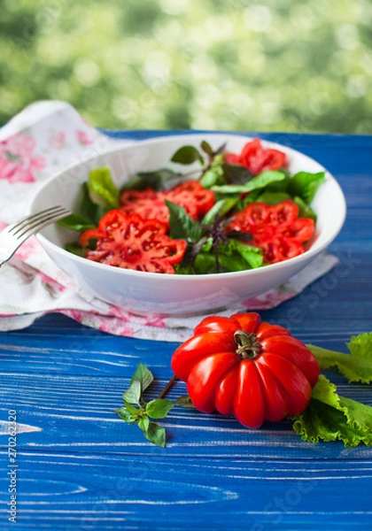 Fototapeta Salad with large ripe red tomato with basil on a blue wooden table, tomato variety.