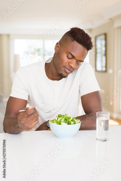 Fototapeta Handsome african young man eating a healthy vegetable salad using a fork to eat lettuce, happy and smiling sitting on the table