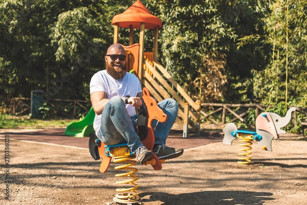 Obraz Playful crazy man (dad) riding wooden rocking horse in a park - adult guy (hipster with beard) having fun on a playground
