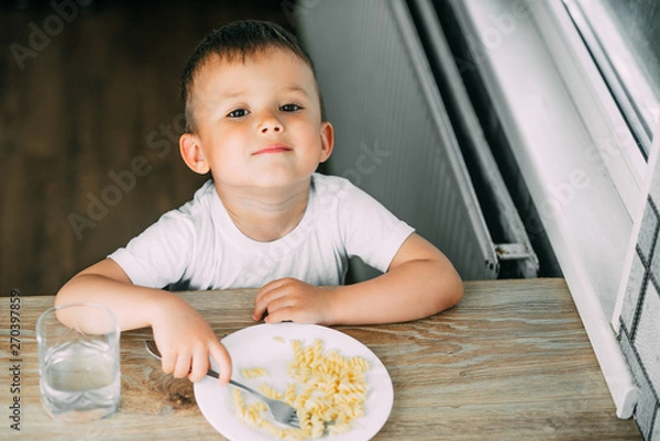 Fototapeta a little boy eats pasta in the form of a spiral in the afternoon in the kitchen on their own