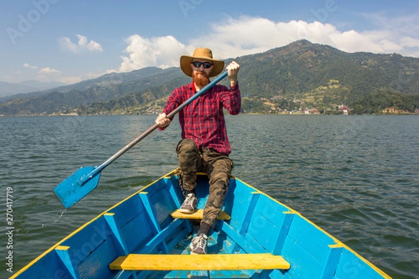 Fototapeta bearded man in a red shirt, hat and sunglasses in a blue wooden boat with a paddle in his hands, on the lake against the background of green mountains