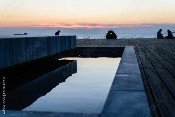 Fototapeta People sitting in the pier in Thessaloniki, Greece. Watching the sunset and talking to each other