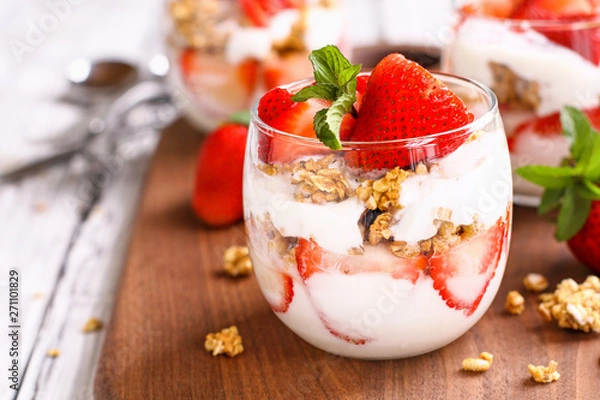 Fototapeta Zdrowe śniadanie składające się z truskawkowych parfaitów ze świeżych owoców, jogurtu i muesli na rustykalnym białym stole. Niewielka głębokość pola, z selektywnym naciskiem na szklany słoik z przodu. Niewyraźne tło.