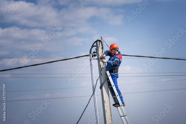 Fototapeta electrical engineer performs wiring on a high pole standing on the stairs. high-rise electrical work. podkluchenie house to the power lines