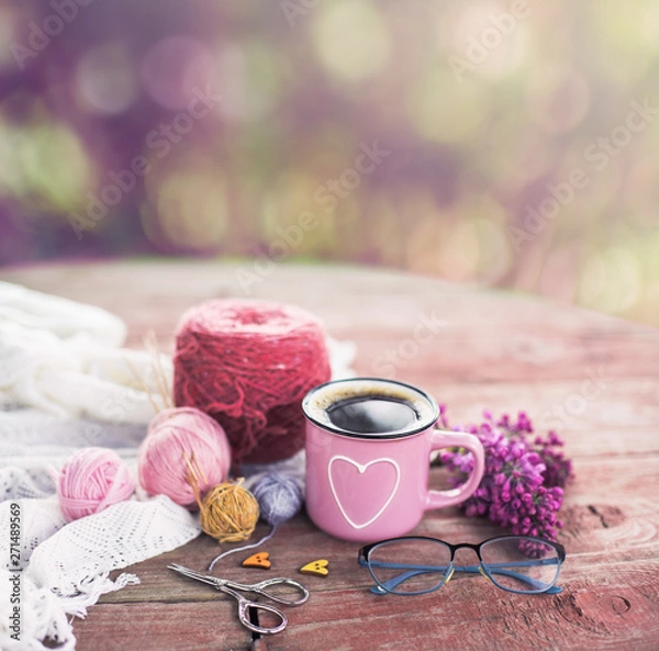 Fototapeta  Women's hobby. Crochet and knitting. Working space. Bouquet of lilac in a cup, balls of yarn, needles, scissors, knitwear, crochet hooks on the wood table  in the cozy home. 