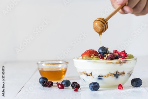 Obraz Plain yogurt with strawberry, blueberries, kiwi, granola, pomegranate in a glass bowl and honey on white wooden texture, healthy food and plant-based food concept