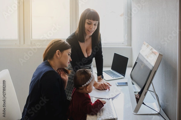 Fototapeta close up photo of two women in office outfit working on computers and discussing a report on the floor; one of them is with a baby girl in her arms