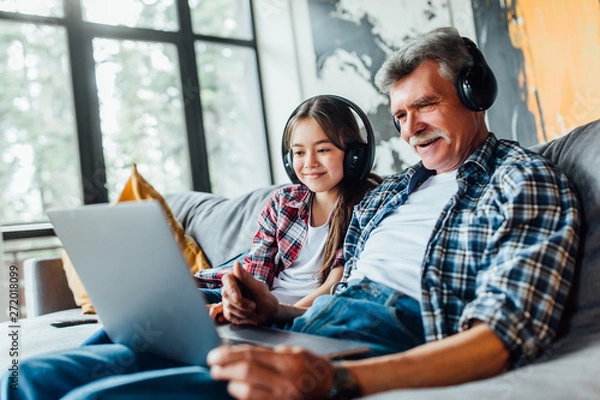 Fototapeta Cute grandchild and her grandfather listening music on digital tablet while sitting on sofa.