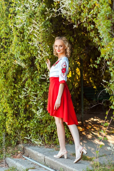 Fototapeta full length portrait of attractive woman in stylish red white dress posing near floral arch in garden. standing and looking at camera with smile. outdoor shot in the park at summer daytime.