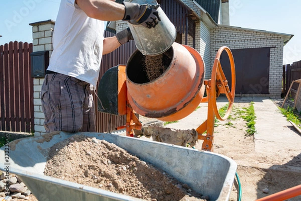 Obraz builder working with shovel during concrete cement solution mortar preparation. construction worker with a bucket in his hands loads a concrete mixer.orange concrete mixer prepares cement mortar