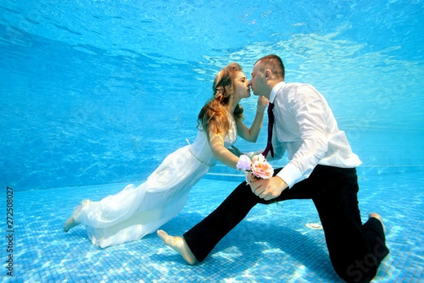 Obraz Happy bride and groom hold hands and kiss underwater at the bottom of the pool on a Sunny day. Wedding bouquet in hand. Portrait. Concept. Underwater photography. Horizontal view