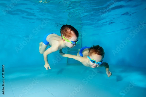 Obraz Two beautiful sporty little boys dive to the bottom of the swimming pool, on a blue background. View from the bottom of the pool. Portrait. Underwater photography. Horizontal view