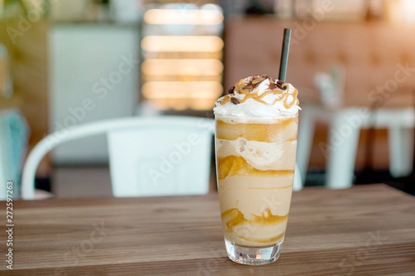 Fototapeta Iced frappe with whipped cream and caramel syrup, with straws and grains of coffee in a tall glass on the wooden table, cold coffee drink