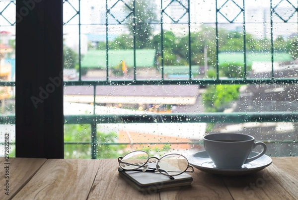 Fototapeta White cup of coffee with steam over a coffee cup and glasses, White cup of caffe americano on wooden table near windows in rainy morning day