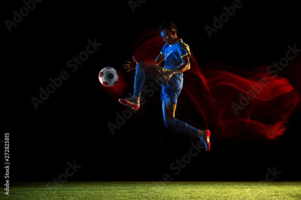 Fototapeta Young african-american male football or soccer player in sportwear and boots kicking ball for the goal in mixed light on dark background. Concept of healthy lifestyle, professional sport, hobby.
