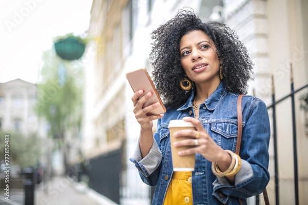 Fototapeta Beautiful girl walking in the street, checking her phone and drinking a coffee