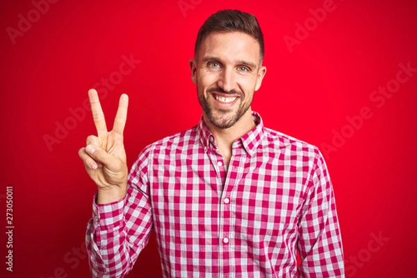 Fototapeta Young handsome man over red isolated background smiling looking to the camera showing fingers doing victory sign. Number two.