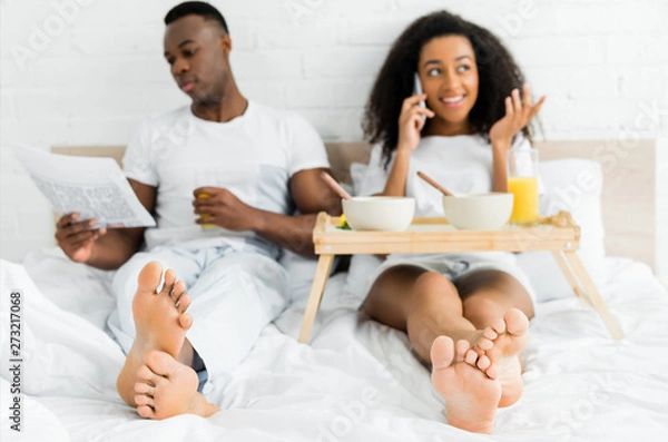 Fototapeta selective focus of african american couple lying on bed, using smartphone and reading newspaper