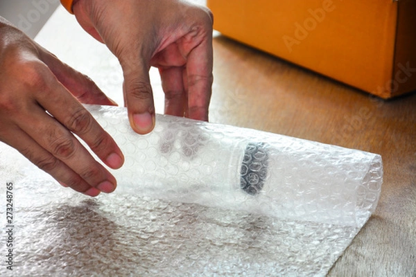 Obraz close up of a guy's hands using bubble wrap to pack a glass bottle before put into cardboard box deliver to customer, e commerce shipping, online business trading concept