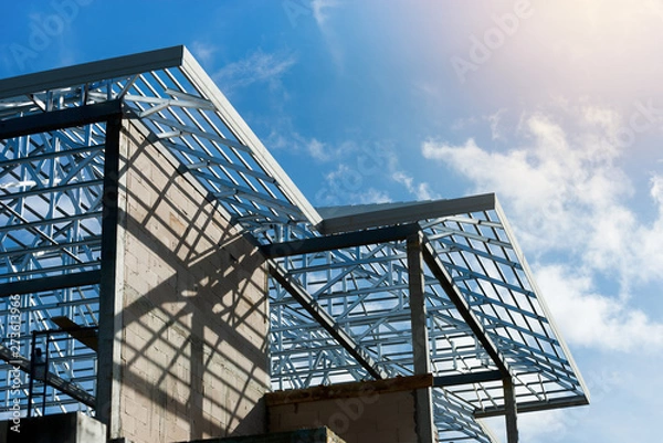 Fototapeta Roof frame of under construction house..Metal roof frame structure of a two story house without  roof tiles under sunny cloud blue sky,low angle view.