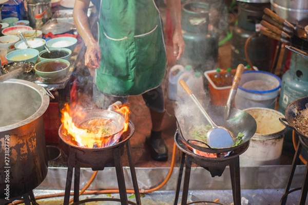 Obraz Chef fries in a wok on the vast flame of the stove with rising steam at a street cafe in an alley adjacent to Yaowarat Road, the main artery of Chinatown of Bangkok, Thailand famous with street food