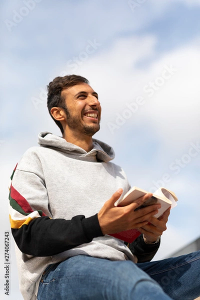 Fototapeta Young Bearded Man Reading A Book Sitting In A Park During The Morning.