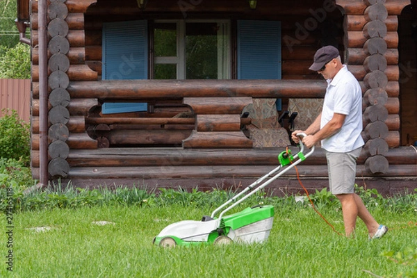Fototapeta A man mows the grass with a lawn mower in the backyard