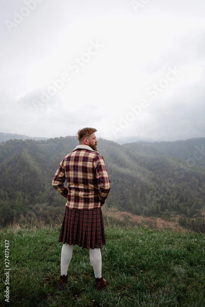 Fototapeta man kilt with mustache beard standing on the top Green Mountain grass, dressed plaid shirt, white socks brown shoes, looks around. it is snowing from the sky. fog around.Caucasian
