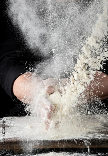 Fototapeta chef in black uniform sprinkles white wheat flour in different directions