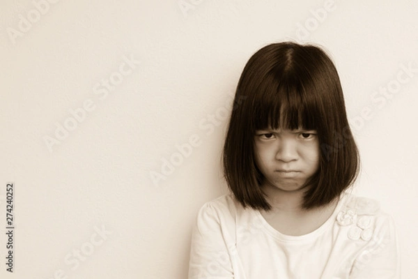 Fototapeta Angry little Asia kid in white shirt in cream wall background. Anger Asian student girl express her emotion on her face. Spoiled child does not get what she want. Disobedience Day