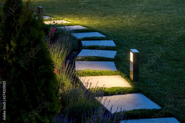 Obraz marble path of square tiles illuminated by a lantern glowing with a warm light in a backyard garden with a flower bed and a lawn copy space.