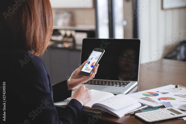 Fototapeta Business people are holding a smartphone and use it to check information at hand ,Businesswomen work in office using a laptop computer, job analysis and job description checking