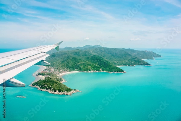 Fototapeta Aerial view of airplane flying above   clouds and sky. Airplane wing fly over tropical green island. View from the plane window.