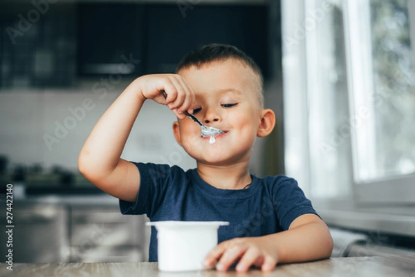 Fototapeta Cute boy in the kitchen eating yogurt from a white container for yogurt, there is a place for advertising
