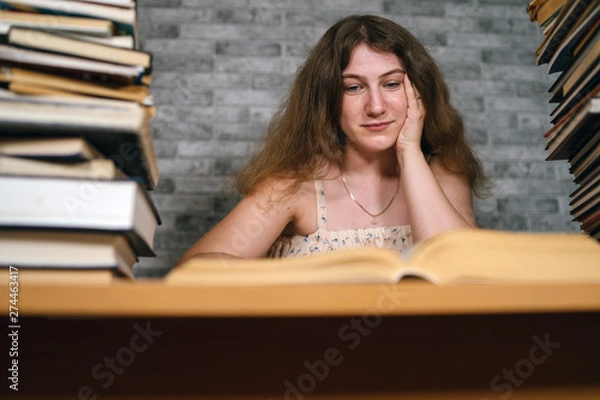 Fototapeta Tired female student reading among books. Pensive young woman sitting at table with pile of book and reading on background of grey wall