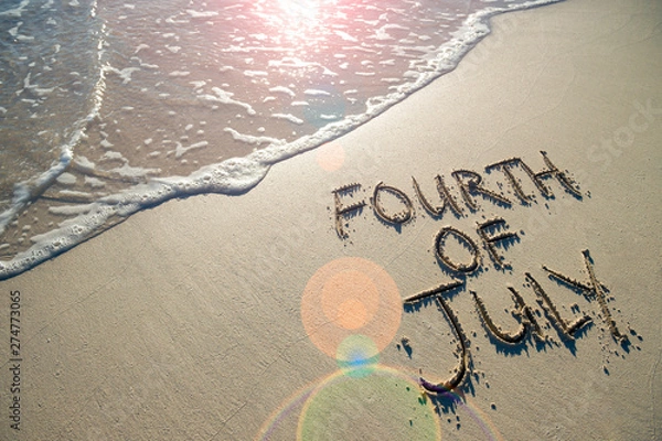 Fototapeta Fourth of July message written for the American Independence Day holiday in smooth sand with sunny lens flare view of an incoming wave on the beach