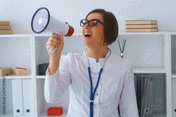 Obraz young beautiful doctor in white gown stay in her consulting room hold loudspeaker
