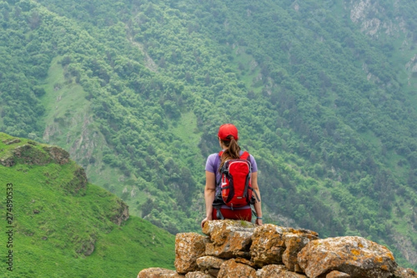 Fototapeta View from the back, a girl with a backpack in a red cap sits and admires the view of the mountains. Travel concept