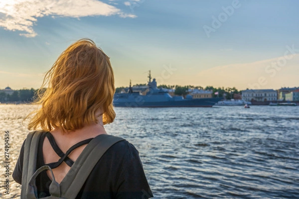 Fototapeta Redhead tween tourist girl on Neva river embankment looking at warships and architectural ensemble in summer evening at sunset. Authentic lifestyle moments, travel concept. Saint Petersburg, Russia