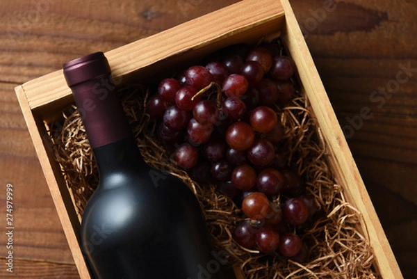 Fototapeta A high angle closeup of a red wine bottle in a wood crate with grapes.