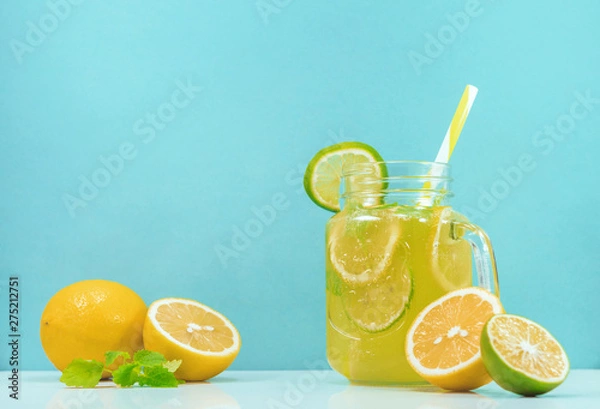 Fototapeta Lemonade drink of soda water, citrus and mint on the light blue background. Cocktail glass with handle and straw, sliced lemon and lime, front view